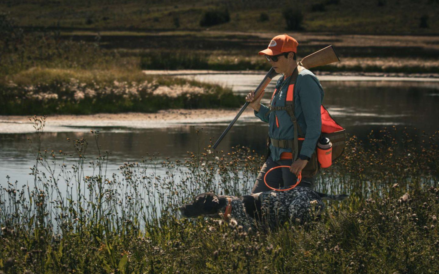 person carrying a shotgun walking with a dog next to a river