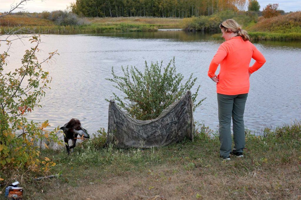 a woman in a bright orange top standing in front of a pond watching a dog come out of the water