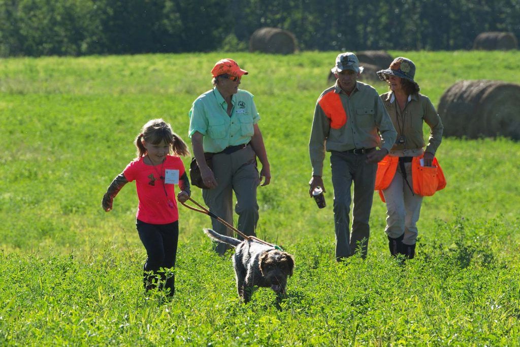 a young girl running through a field with a dog on a leash followed by three people