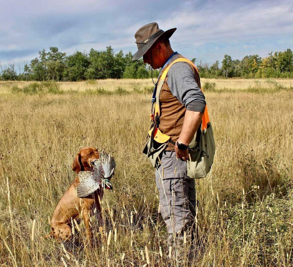 a man in hunting gear looking down at a dog with a duck in it's mouth