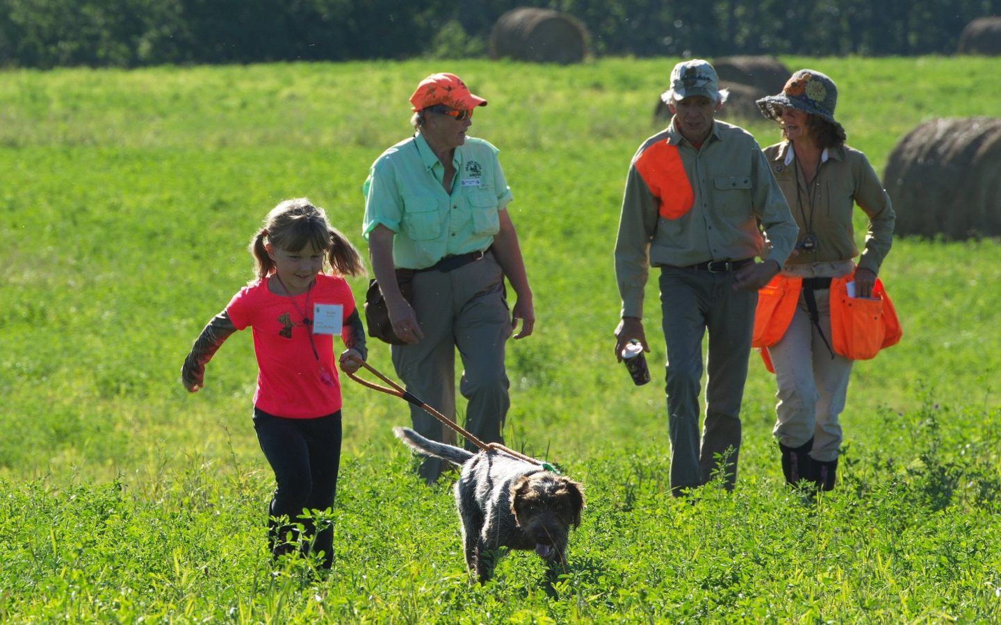 a young girl running through a field with a dog on a leash followed by three people